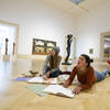 Two students drawing in their sketchbooks on the floor of a museum
